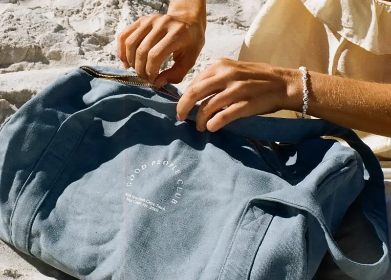 Hände öffnen eine blaue Tasche am Strand, am Handgelenk ein Mon Coeur Armband